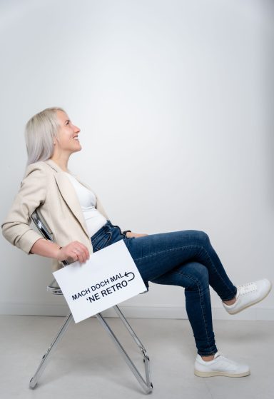 Frau mit langen, blonden Haaren sitzt auf einem Stuhl und hält ein Schild.