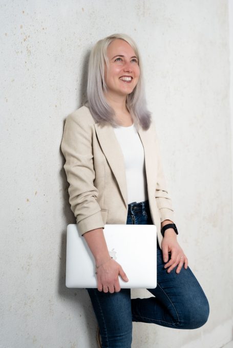 Junge Frau mit blonden Haaren, die einen Laptop hält, lächelt vor einer neutralen Wand.