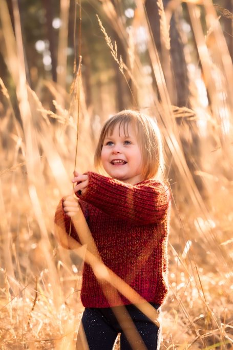 Ein kleines Kind mit einem roten Pullover steht in hohem, goldenem Gras und lächelt.