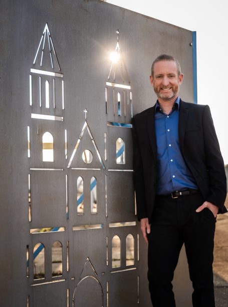 Mann in Anzug steht neben einer geschlossenen Stahlwand mit einem historischen Gebäudemotiv.
