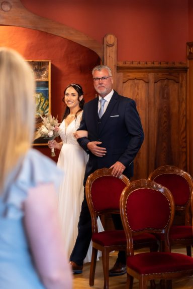 Eine Braut in einem weißen Kleid steht neben ihrem Vater in einem festlichen Raum.