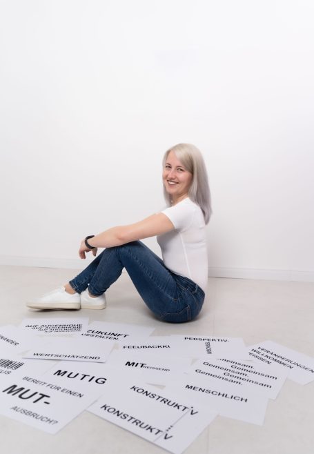 Junge Frau mit grauen Haaren sitzt auf dem Boden umgeben von beschrifteten Papierblättern.