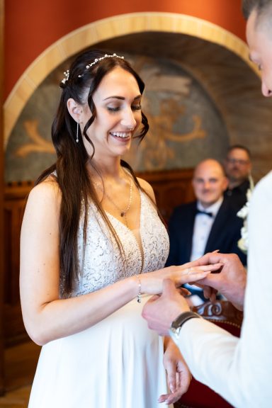 Eine Braut steckt ihrem Bräutigam den Ring an, im Hintergrund ein lächelnder Mann.