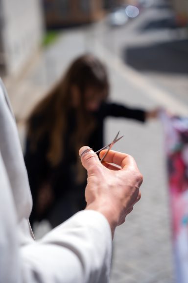Eine Hand hält eine Schere, im Hintergrund eine unscharfe Person, die etwas betrachtet.
