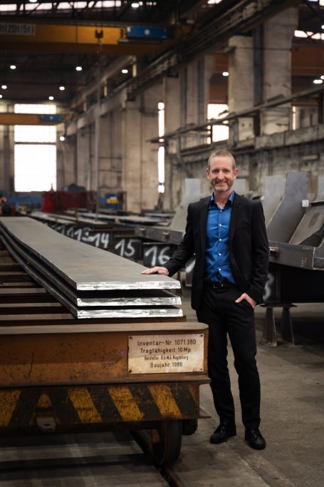 Mann in Anzug steht neben gestapelten Metallplatten in einer Fabrikhalle.