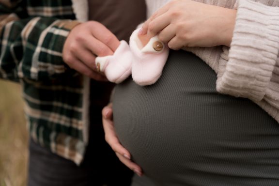 Hände legen rosa Babyschuhe auf einen runden Babybauch im Freien.