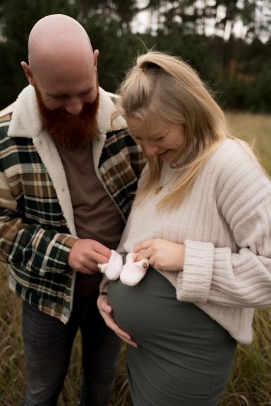 Ein Paar steht im Freien, die Frau hält Babyschuhe über ihrem Babybauch.