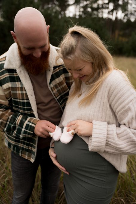 Schwangere Frau mit Partner, die kleine Babyschuhe hält, umgeben von Natur.