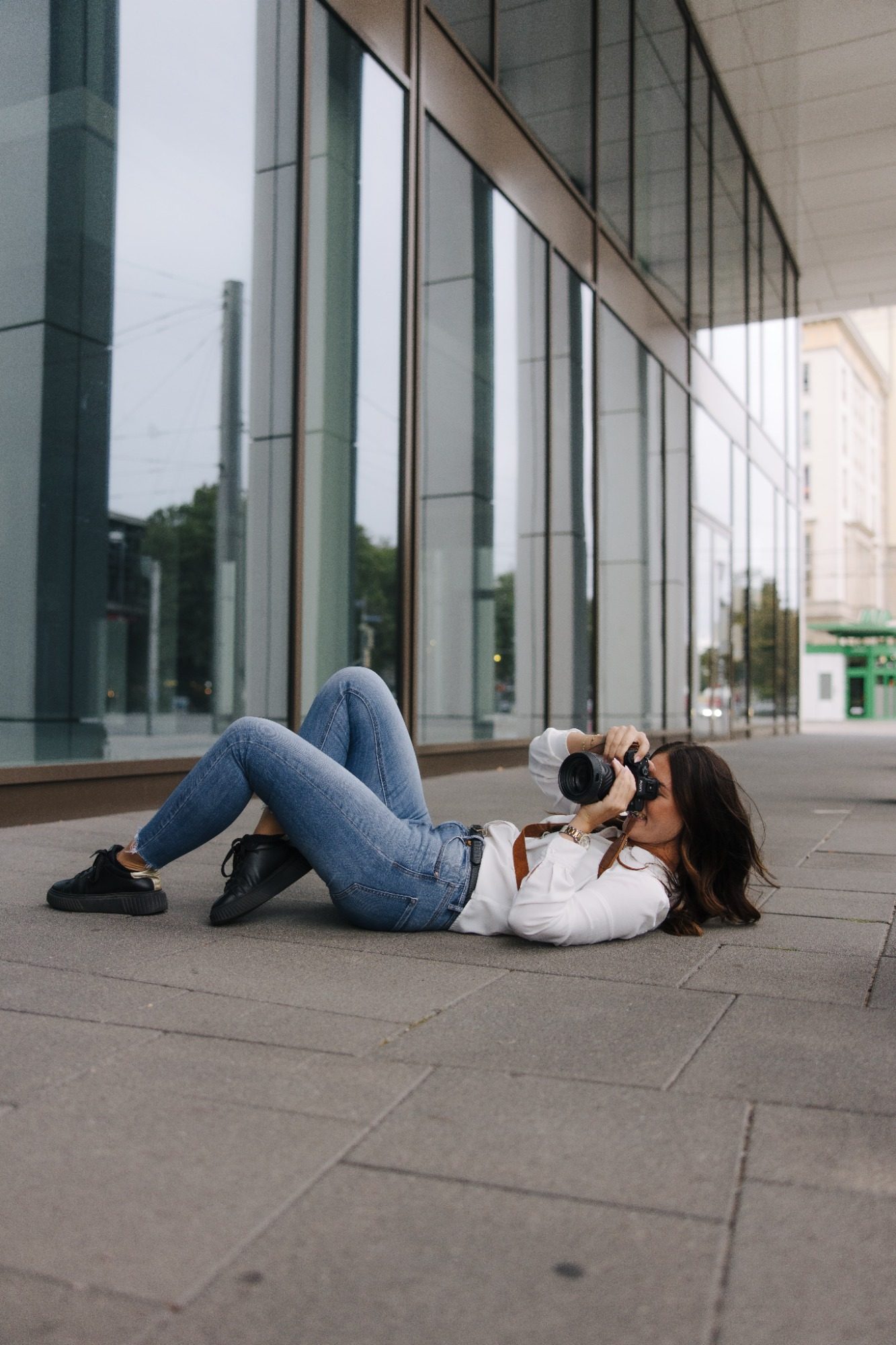 Frau liegt auf dem Boden und fotografiert mit einer Kamera, moderne Architektur im Hintergrund.