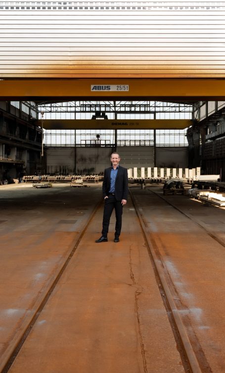 Mann in Anzug steht in einer industriellen Halle mit rostigem Boden und hohen Wänden.