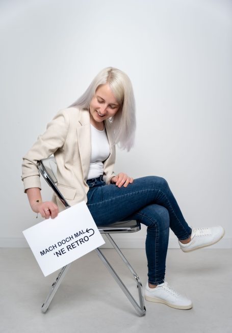 Junge Frau mit blonden Haaren sitzt auf einem Stuhl und hält ein Schild in der Hand.