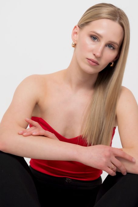 Junge Frau mit langen blonden Haaren in rotem Oberteil sitzt nachdenklich.