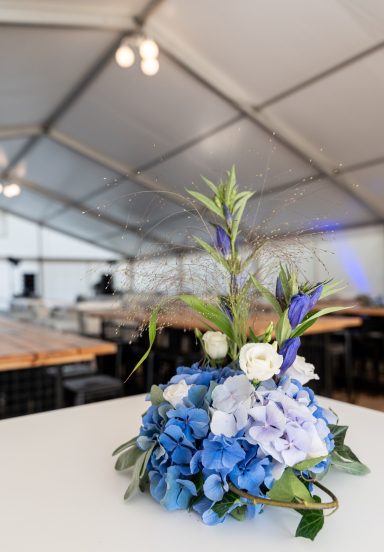 Blumenarrangement mit blauen und weißen Blumen auf einem Tisch in einem Zelt.