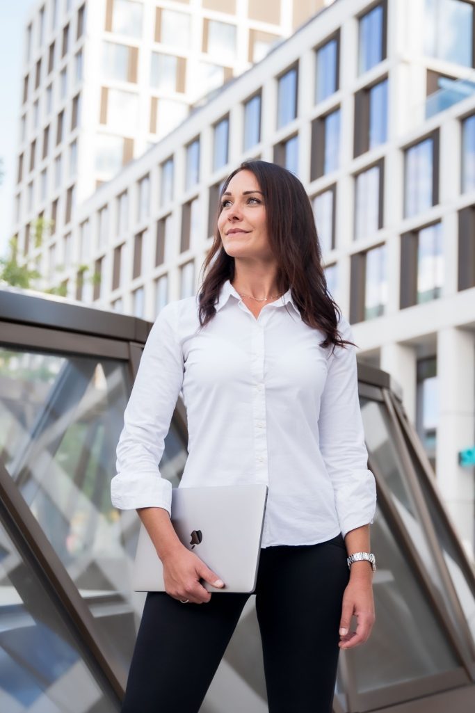 Frau in weißem Hemd hält ein Laptop, steht vor moderner Architektur im Freien.