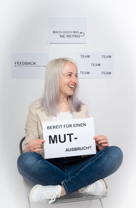Junge Frau mit blonden Haaren sitzt und hält ein Schild mit der Aufschrift „Mut“.