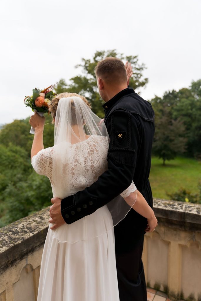 Hochzeitspaar von hinten, Braut mit Schleier und Strauß, Bräutigam umarmt sie.