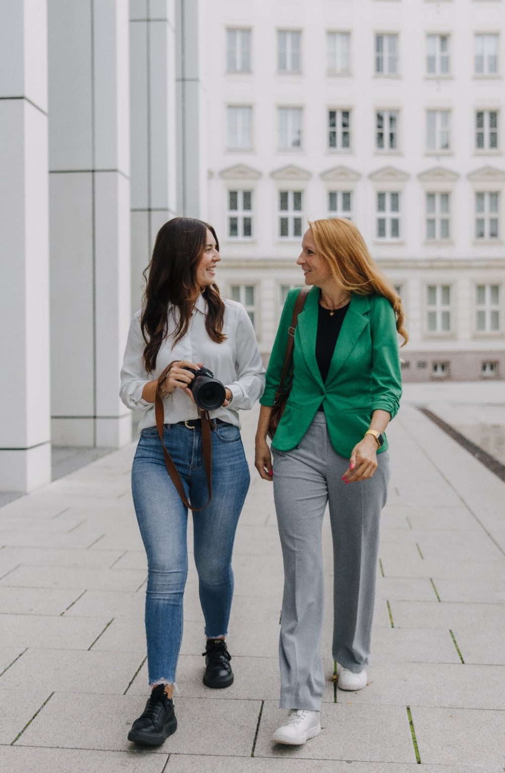 Zwei Frauen gehen in Businesskleidung auf einer städtischen Straße und lächeln sich an.