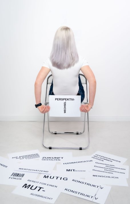 Person mit blonden Haaren sitzt rückwärts auf einem Stuhl, umgeben von beschrifteten Blättern.