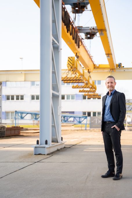 Mann in Anzug steht neben einem Kran auf einem Industriegelände.