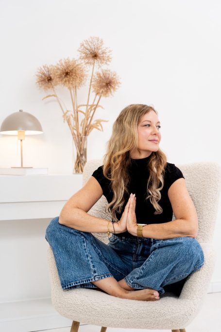 Frau im schwarzen Oberteil sitzt im Sessel, meditiert mit gekreuzten Beinen.
