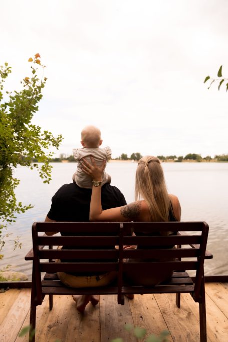 Eltern sitzen auf einer Bank am Wasser, ein Baby wird auf den Schultern des Vaters gehalten.