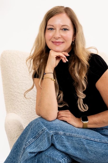 Junge Frau mit langen, blonden Haaren, lächelt lässig auf einem Sessel.