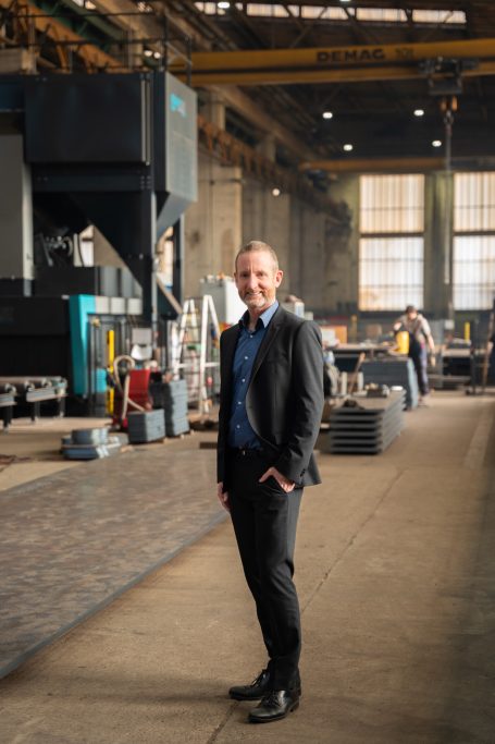Mann in Anzug steht in einer industriellen Umgebung mit Maschinen im Hintergrund.