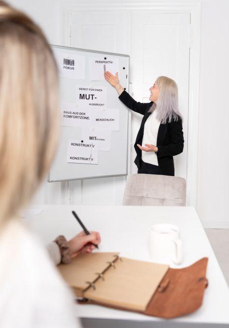 Frau präsentiert vor Flipchart, während eine andere Person notiert.