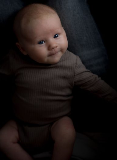 Ein lächelndes Baby sitzt in einem dunklen Hintergrund, trägt ein braunes Oberteil.