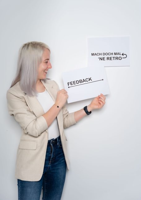 Frau mit kurzer, blonder Haarfarbe hält Schilder mit den Aufschriften „Feedback“ und „Was denkst du?“