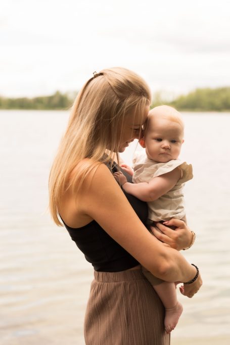 Eine Frau mit langen Haaren hält ein Baby am Ufer eines Gewässers.