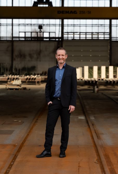 Mann in Anzug steht in einer Industriehalle mit großen Fenstern.