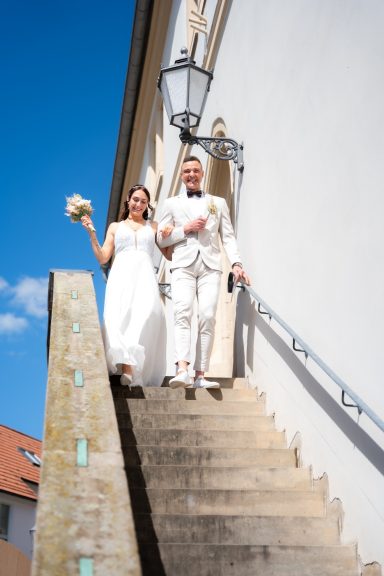 Brautpaar in festlicher Kleidung, das eine Treppe hinuntergeht, mit Strauß in der Hand.