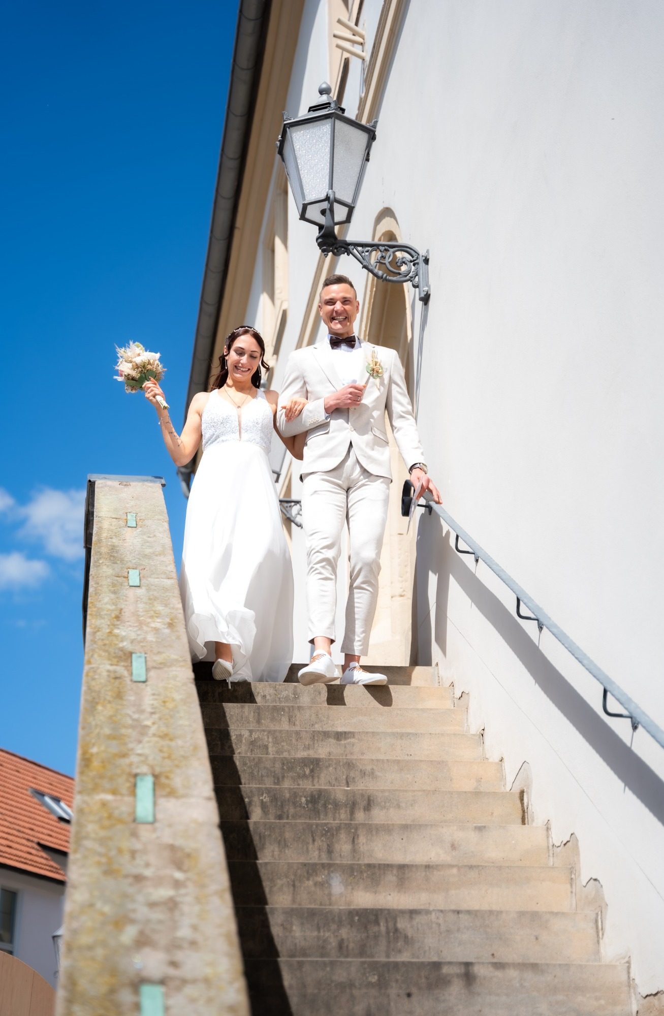 Ein Brautpaar in eleganter Kleidung, das eine Treppe hinuntergeht, mit Blumen in der Hand.