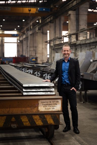 Mann in Anzug steht neben großen Stahlplatten in einer Fabrikhalle.