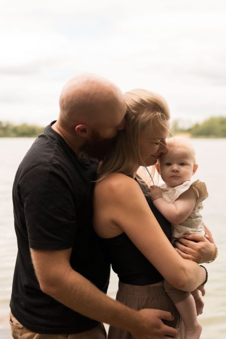 Familie posiert am Wasser, Eltern umarmen sich mit einem Baby in den Armen.