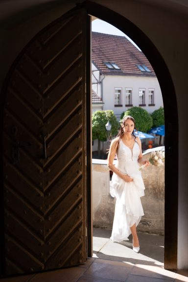Eine Braut steht in einem Türrahmen, im Hintergrund historische Gebäude und Bäume.