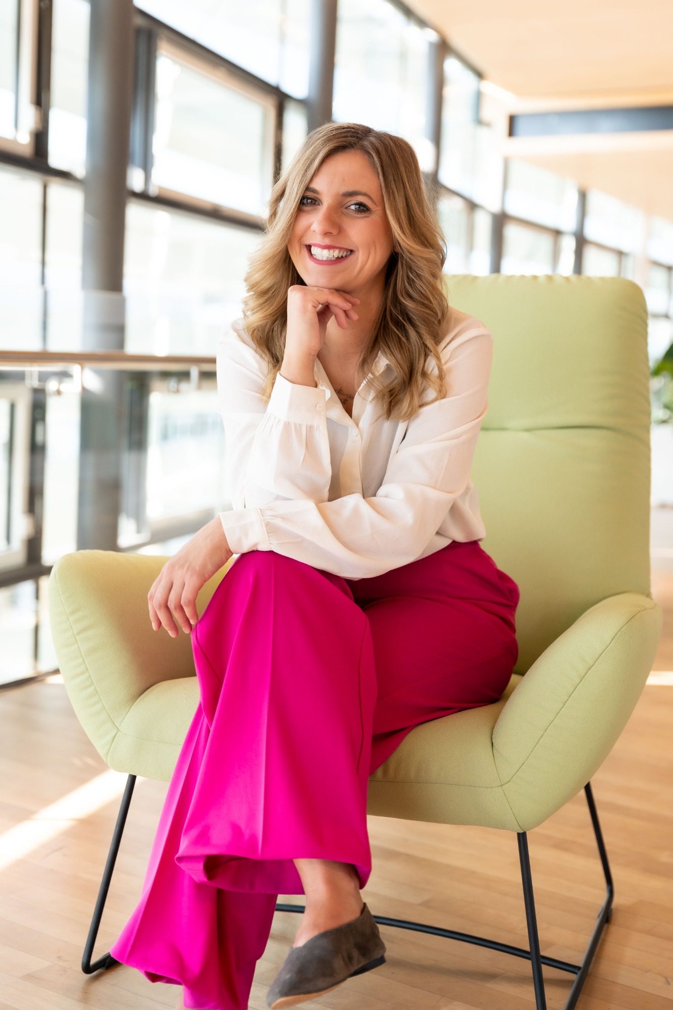 Frau mit blonden Locken in einem weißen Hemd und pinker Hose, sitzt entspannt auf einem Stuhl.