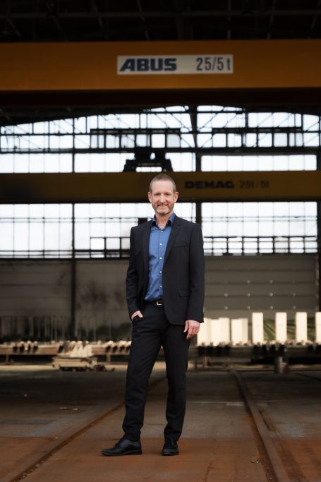 Mann in Anzug steht in einer Industriehalle mit Kränen im Hintergrund.