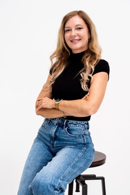 Lächelnde Frau mit langen Haaren, trägt ein schwarzes Oberteil und Jeans.